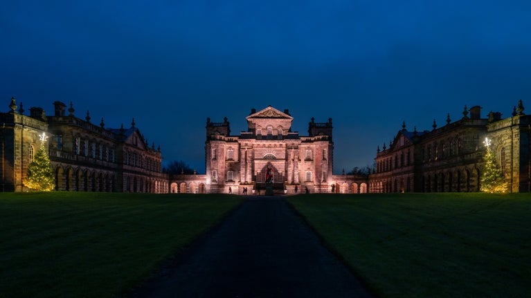 A large stone building flanked by two wings on each side. The central building is lit up in a pale pink colour. Behind the building the sky is a deep navy blue. In the foreground there is a central driveway with grass either side.